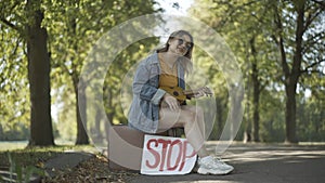 Wide shot of positive hippie woman sitting on suitcase with Stop sign, playing ukulele, and singing. Portrait of relaxed