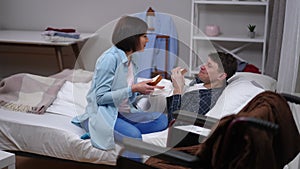 Wide shot positive husband and wife eating sandwiches talking smiling. Ill Caucasian man lying in bed chatting with