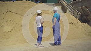 Wide shot of professional female inspector and male worker standing on manufacturing site in front of enormous sand pile