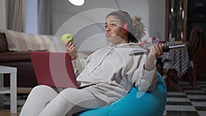 Wide shot of relaxed African American woman eating apple lifting dumbbell sitting with laptop on bag chair. Portrait of