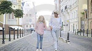 Wide shot of relaxed happy grandmother and granddaughter holding hands and talking as strolling outdoors on sunny day