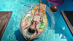 Wide shot of relaxed little girl lying on swimming mattress in blue water putting hands behind head in slow motion