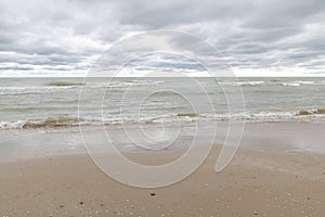 A wide shot of the sandy shore with gentle waves crashing against it, under an overcast sky with clouds in shades of