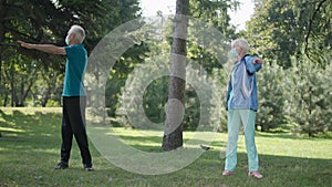 Wide shot senior couple in coronavirus face masks exercising in park warming up in slow motion. Caucasian man and woman