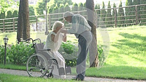 Wide shot of senior man helping beautiful woman to stand up from wheelchair in sunny summer park. Loving husband