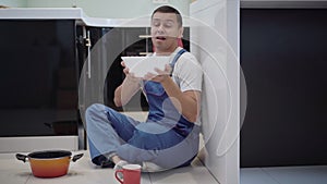 Wide shot of service man in blue uniform sitting on kitchen floor catching water leaking from ceiling. Young Caucasian