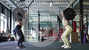Wide shot side view of two confident strong women squatting lifting heavy barbells in gym indoors. Athletic motivated