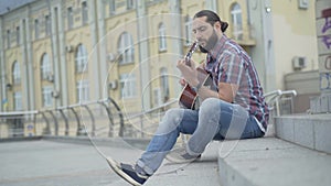 Wide shot side view of young Caucasian man sitting on urban stairs and playing guitar. Handsome bearded musician
