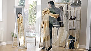 Wide shot of slim glamorous Caucasian lady holding fur coat looking in mirror. Side view of confident young woman