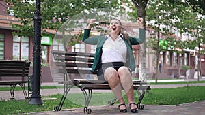 Wide shot successful happy obese businesswoman sitting on bench gesturing victory in slow motion. Portrait of excited