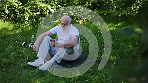 Wide shot thoughtful senior sportsman sitting on exercise mat in summer park scrolling social media in smartphone app