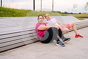 Wide shot of tired obese young woman sitting by bench after intense exercise with personal trainer outdoor in city park.