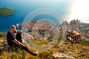 Wide shot from the top of the Srd mountain, man sitting on a rock observing the area around the city of Dubrovnik, orange cablecar