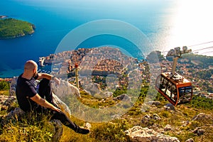 Wide shot from the top of the Srd mountain, man sitting on a rock observing the area around the city of Dubrovnik, orange cablecar