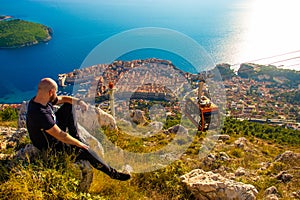 Wide shot from the top of the Srd mountain, man sitting on a rock observing the area around the city of Dubrovnik, orange cablecar