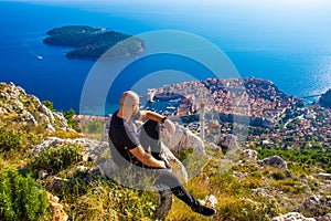 Wide shot from the top of the Srd mountain, man sitting on a rock observing the area around the city of Dubrovnik, orange cablecar