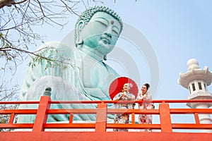 Wide shot of two Asian pretty women stay close to red railing in the area of base of green big buddha statue and they look to left