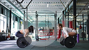 Wide shot two confident athletic sportswomen lifting barbells in slow motion standing up in gym with equipment. Side