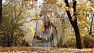 Wide shot young blond woman in yellow raincoat throwing autumn leaves in park