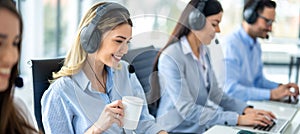 Wide view of beautiful blonde girl with coffee cup working on computer accompanied with group of call center operators.