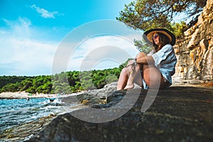 Wider shot of an attractive brunette sitting outside on a rock on a beach in croatia. Amazing summer day on a vacation , bright