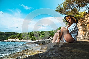 Wider shot of an attractive brunette sitting outside on a rock on a beach in croatia. Amazing summer day on a vacation , bright