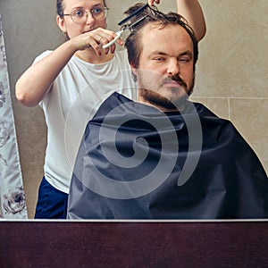 Wife cuts a man`s hair with scissors in front of the mirror, copy space. The concept of unkempt appearance and problems in