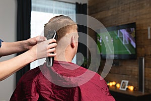 Wife cutting husbands hair at home in front of TV with scissors and holding comb in other hand photo