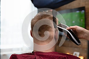 Wife cutting husbands hair at home in front of TV with clipper and holding comb in other hand. Take care each other photo