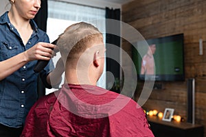 Wife cutting husbands hair at home in front of TV with clipper and holding comb in other hand photo