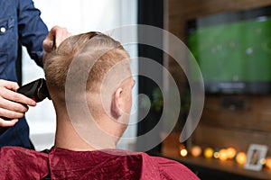 Wife cutting husbands hair at home indoors in front of TV with clipper and holding comb in other hand photo
