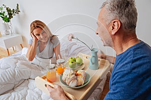 Wife looking at husband holding tray with breakfast and flowers