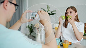 Wife posing with apples while husband taking photo with smartphone in kitchen