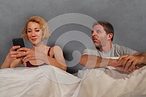 Wife using mobile phone in bed with her angry frustrated husband and the man feeling ignored upset and bored in woman internet add