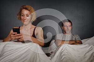 Wife using mobile phone in bed with her angry frustrated husband and the man feeling ignored upset and bored in woman internet add