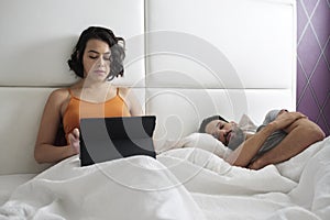 Wife Working In Bed At Home And Husband Sleeping