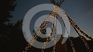 A wigwam made of sticks wrapped with a luminous garland in the evening outdoors