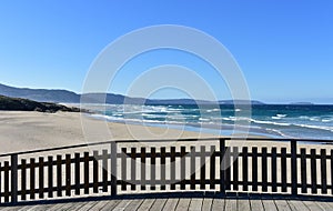 Wild beach with golden sand and wooden boardwalk. Blue sea with waves and foam, sunny day. Galicia, Spain.