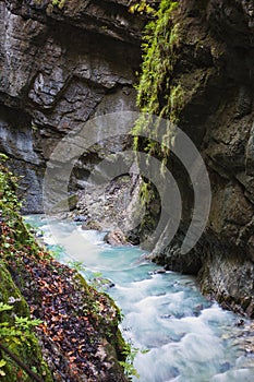 A wild mountain river with clear blue water flows through the open area of the Partnachklamm gorge in Germany. The rapid