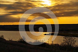 Wild natural landscape at sunset, golden color with dark silhouettes, late autumn, lake, dry grass, bare branches of trees without