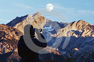 wildlife observer with binoculars, moon above mountain terrain