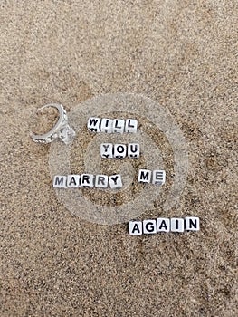 Will you marry me again written on sand at the beach with a beautiful silver engagement ring