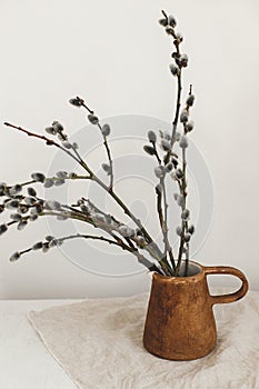 Willow branches in stylish rustic vase on wooden table. Modern Easter still life. Pussy willow branches in home, natural spring