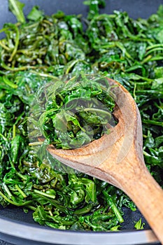 Wilted arugula or rocket salad, preparing a side dish in frying pan, vertical closeup