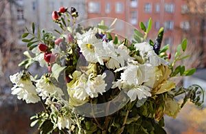 A wilted frozen bouquet of white roses and chrysanthemums, greenery on open window against the background of the street and houses