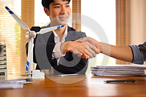 A wind turbine model on meeting table against businesspeople shacking hands on background