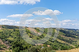 The wind turbines in the top of the grass hill underneath the group of white clouds and blue sky on a sunny day, produced