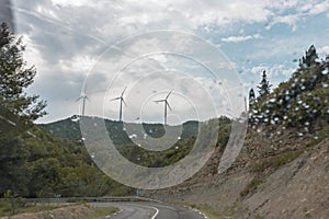 Wind turbines under a cloudy sky seen through the wet glass window of the car