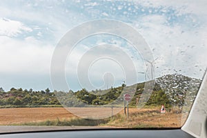 Wind turbines under a cloudy sky seen through the wet glass window of the car