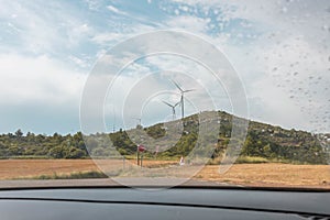 Wind turbines under a cloudy sky seen through the wet glass window of the car